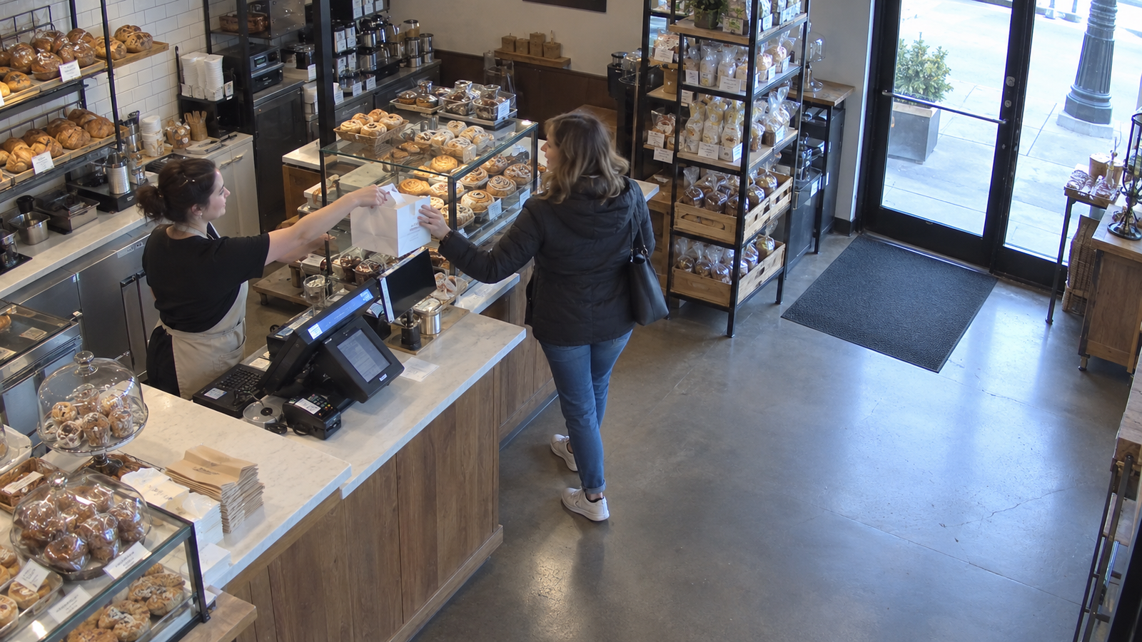 Simulated security camera view of a bakery cashier handing an order to a customer